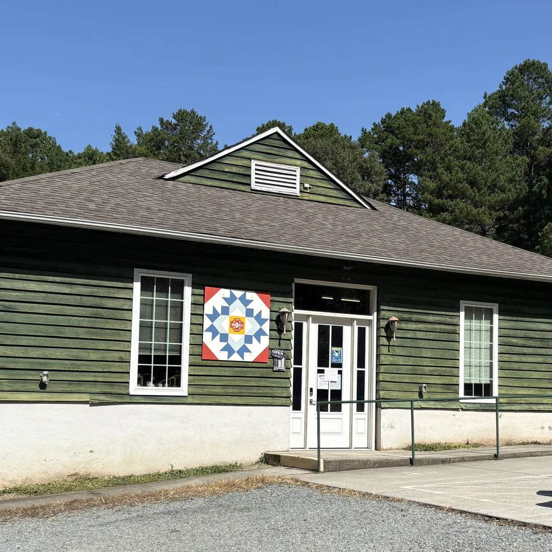 Green storefront building with barn quilt sign and posted hours