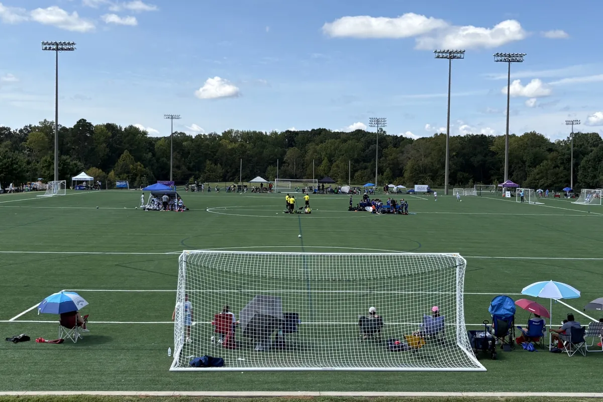 Match in progress on a synthetic turf field at Richard Barry Memorial Park, with spectators along the sideline