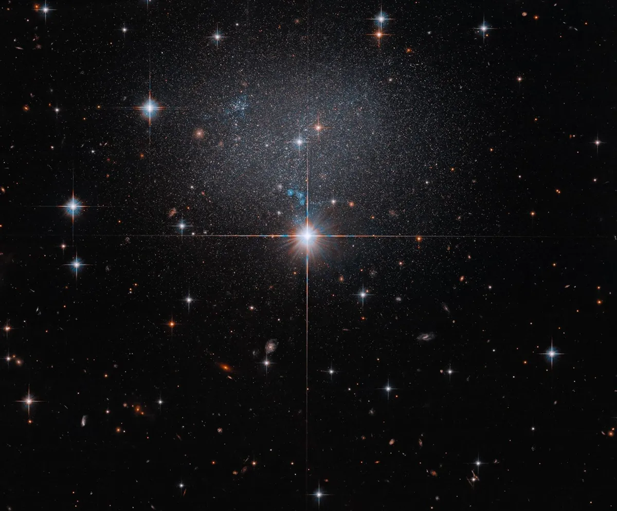 องค์การ NASA เผยภาพถ่ายล่าสุดจากกล้องโทรทรรศน์อวกาศฮับเบิล แสดงให้เห็นกาแล็กซีแคระ ESO 490-017 ที่มีความสว่างพื้นผิวต่ำจนดูเหมือนฝูงดาวที่เลือนลาง ท่ามกลางหมู่กาแล็กซีห่างไกลในอวกาศลึก เพื่อศึกษากระแสคอสมิกและการกระจายตัวของสสารในเอกภพผ่านอิทธิพลของแรงโน้มถ่วง