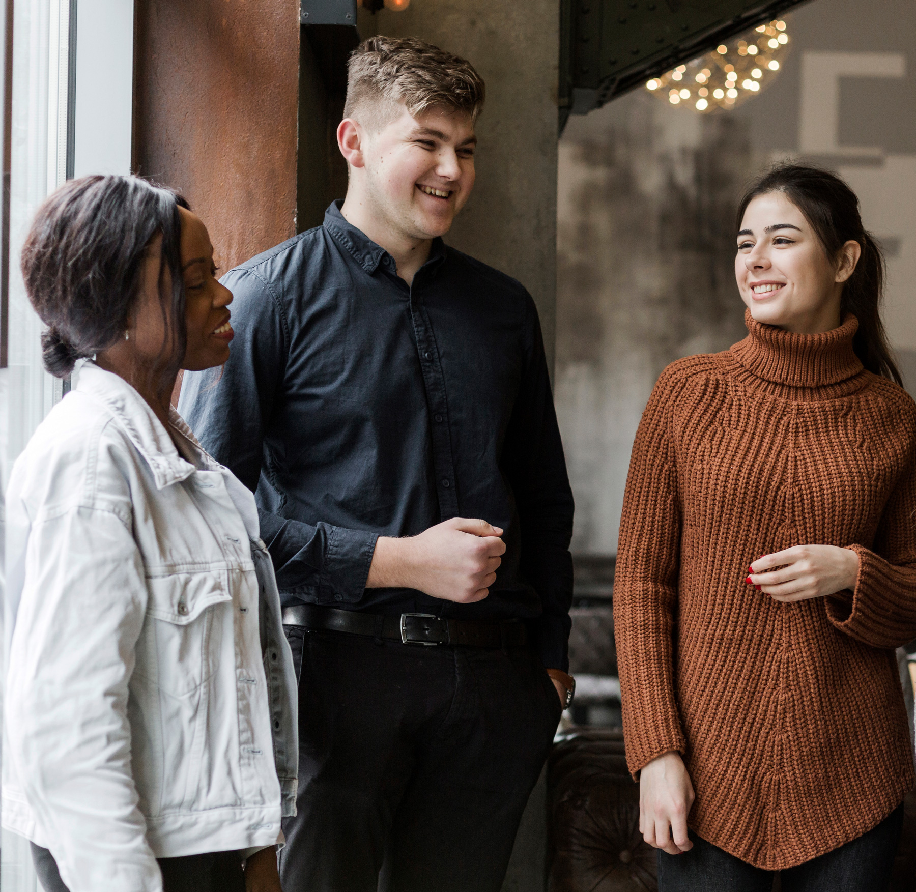 Drei junge Menschen, die lächelnd und im Gespräch an einem Fenster in einem Innenraum stehen.