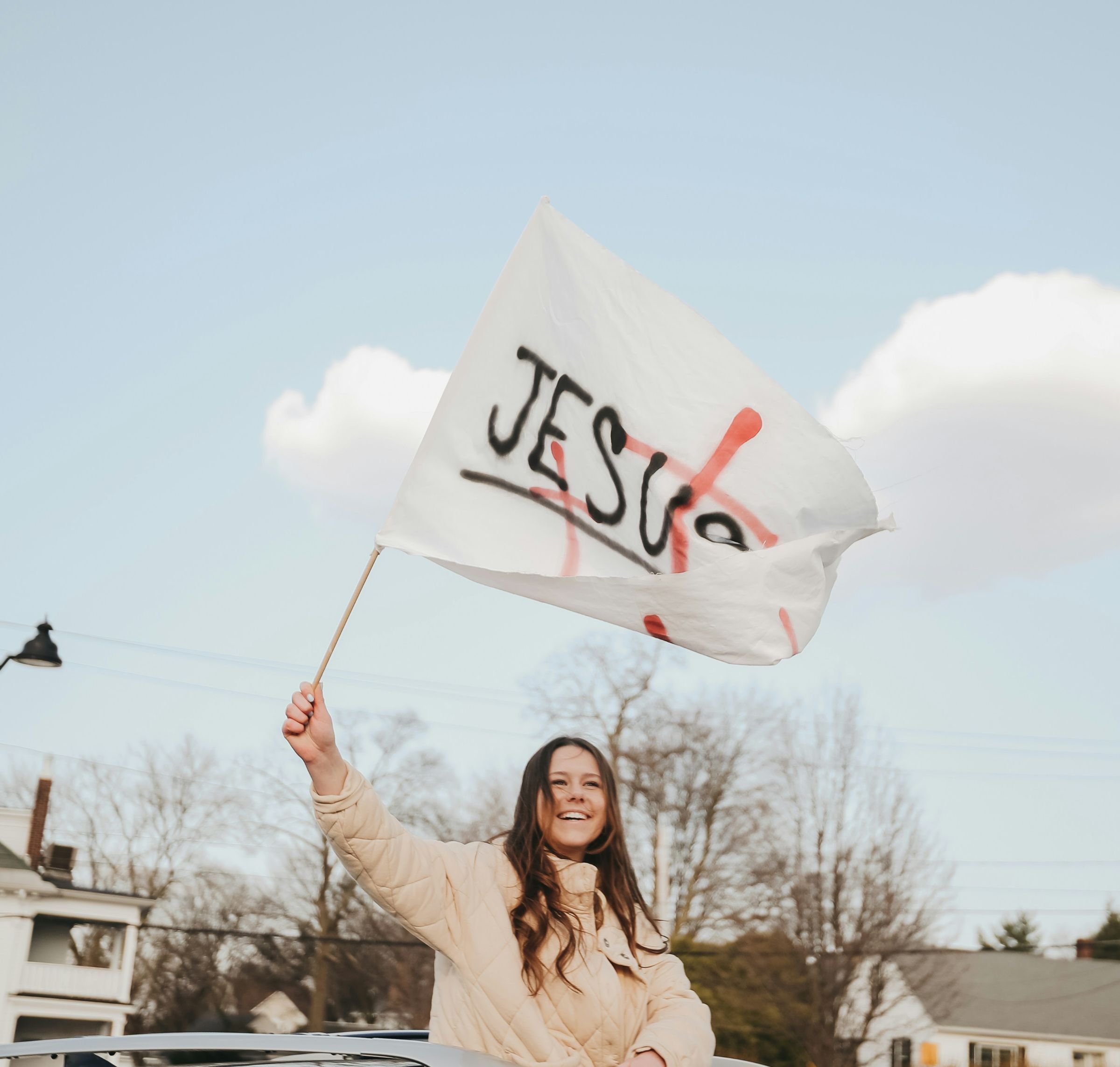 Lächelnde Frau hebt eine weiße Flagge mit der Aufschrift 'JESUS' in der Hand, im Hintergrund Häuser und Bäume.