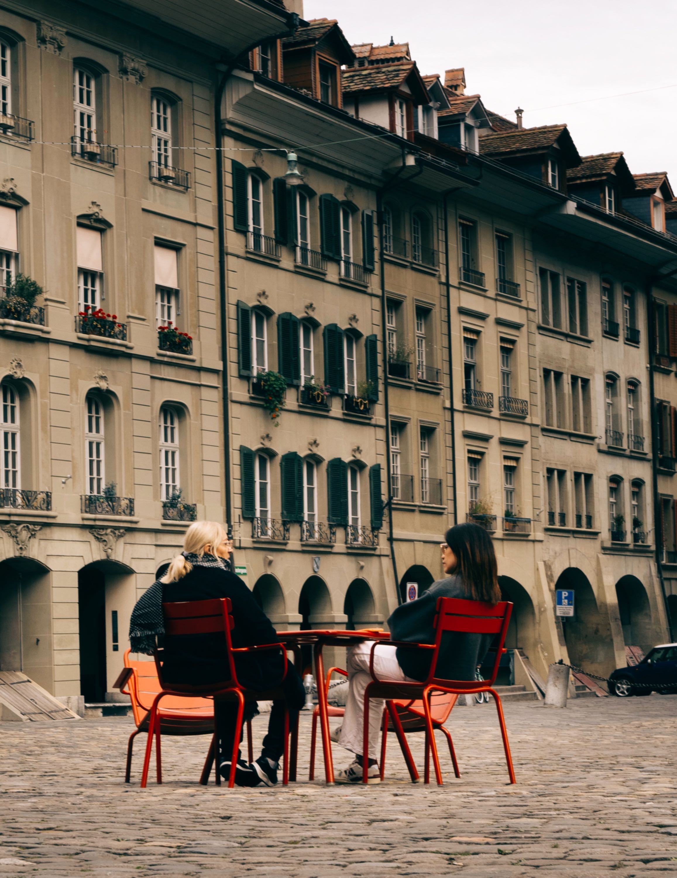 Zwei Frauen sitzen auf roten Stühlen an einem Tisch auf Kopfsteinpflaster vor einem historischen Gebäude.