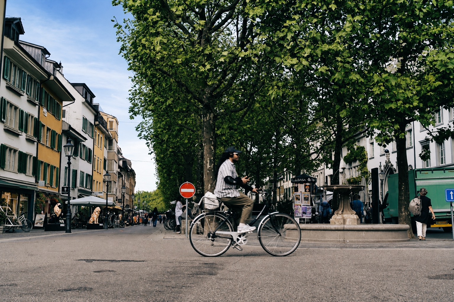 Person fährt Fahrrad an einem Platz mit Bäumen, Gebäuden und einem Brunnen in der Stadt.