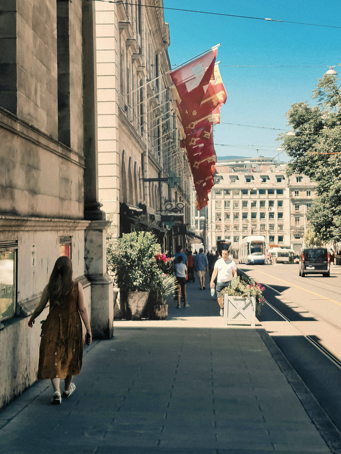 Frau im braunen Kleid geht auf einem Bürgersteig mit roten Fahnen und Straßenbahn im Hintergrund.