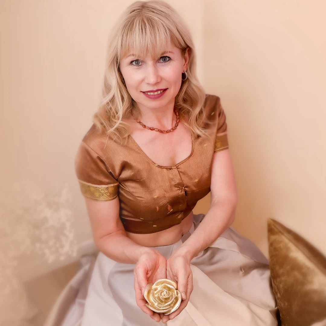 Lächelnde Frau mit blonden Haaren, in brauner Bluse und hellem Rock, hält eine goldene Rosenfigur in den Händen.