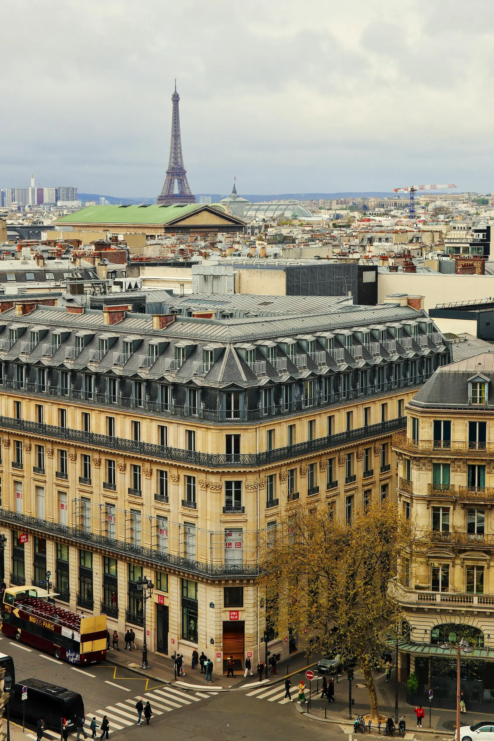 Vue aérienne d'un immeuble haussmannien avec des passants et un bus touristique à Paris, la tour Eiffel en arrière-plan sous un ciel nuageux.