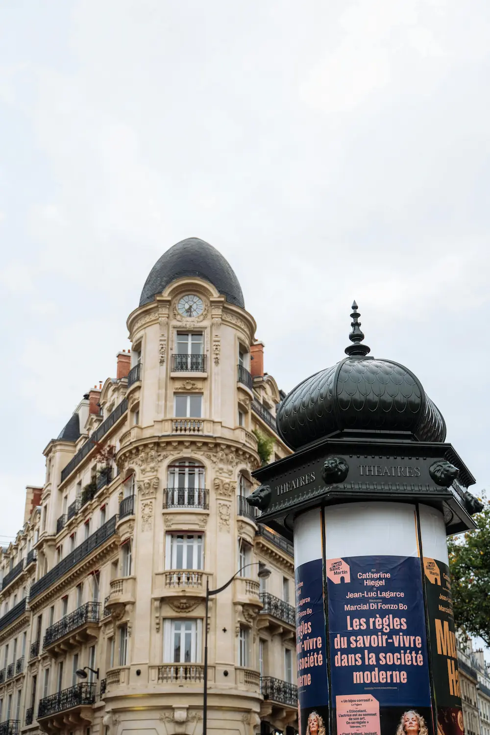 Kiosque d'affichage de théâtre noir orné de têtes de lion devant un immeuble haussmannien avec un toit en forme de clocher à Paris.