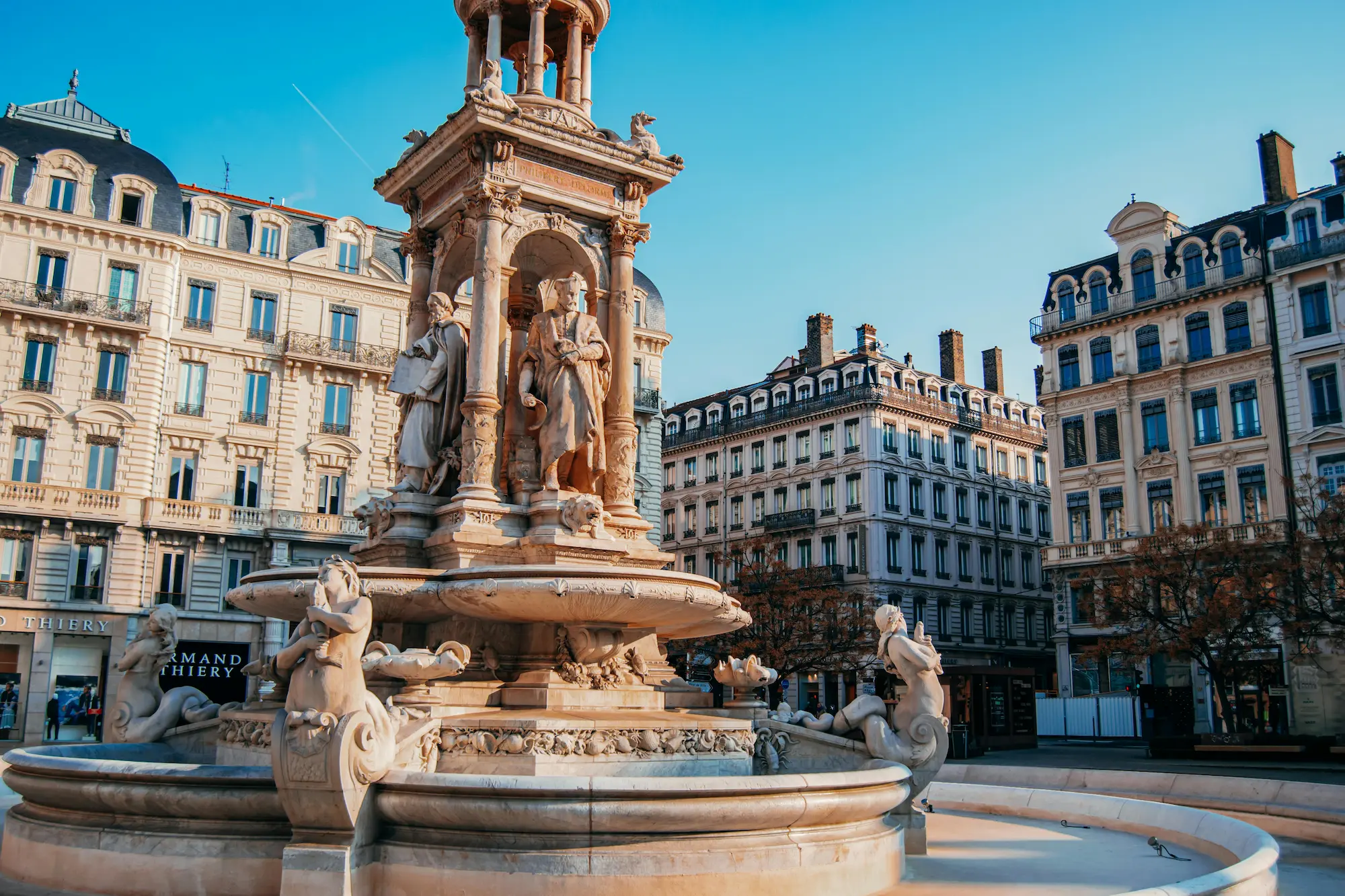Fontaine monumentale en pierre avec statues détaillées, entourée de bâtiments historiques sous un ciel bleu clair.