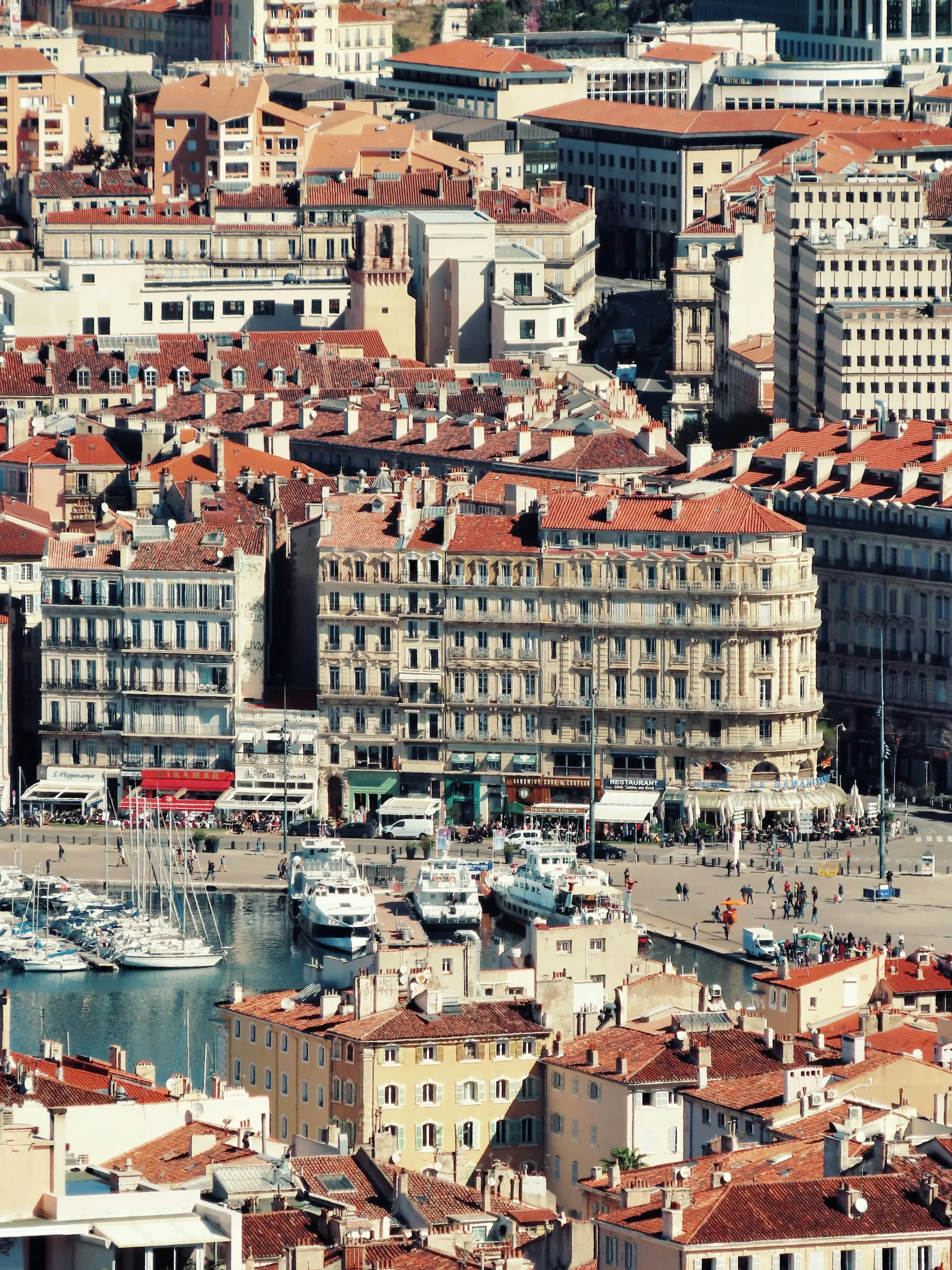 Vue aérienne d'un port avec des yachts et des bateaux amarrés, entouré de bâtiments anciens aux toits en tuiles rouges.