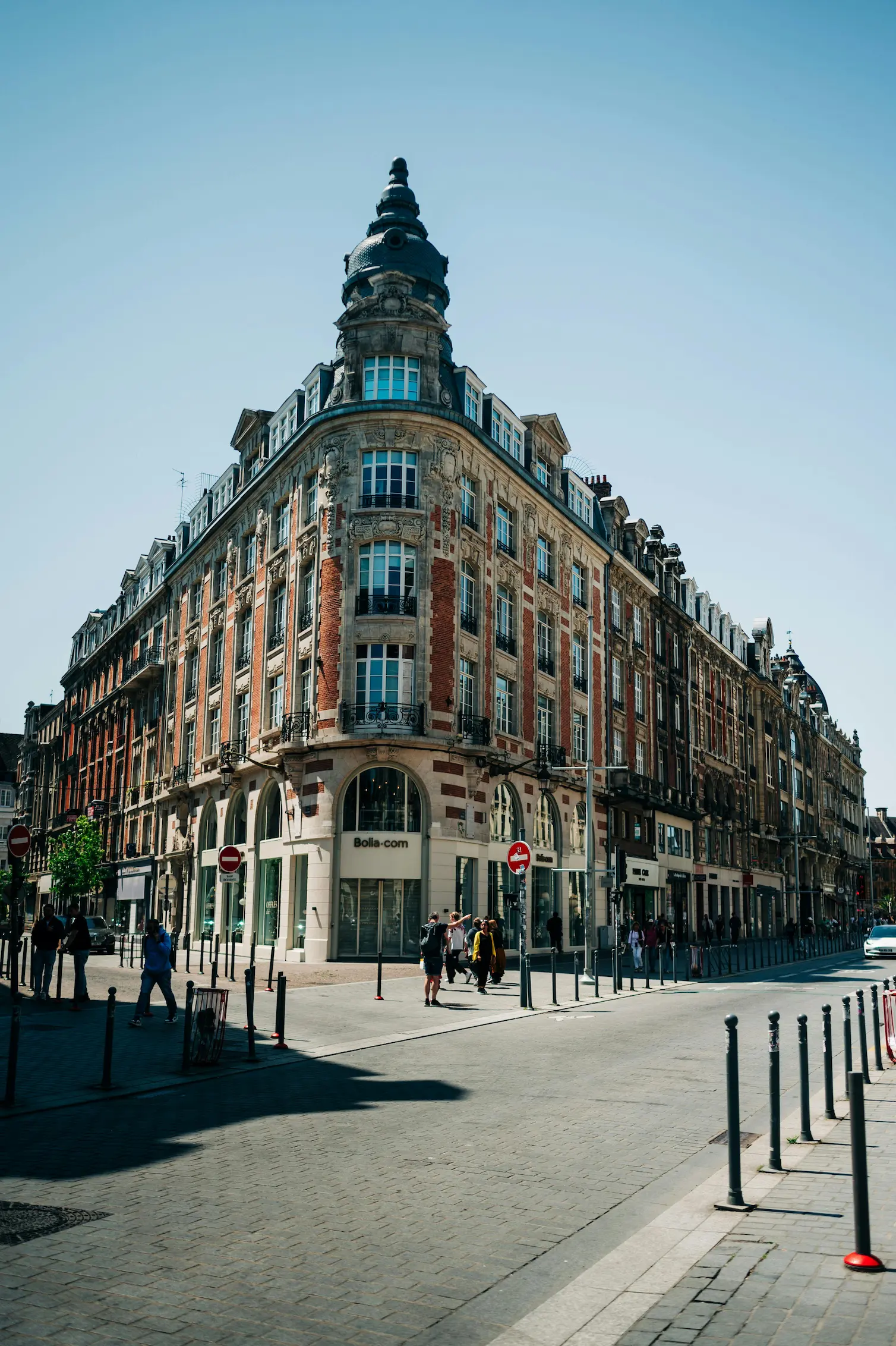 Bâtiment en coin de style classique avec une toiture en dôme, situé au bord d'une rue pavée avec des piétons.