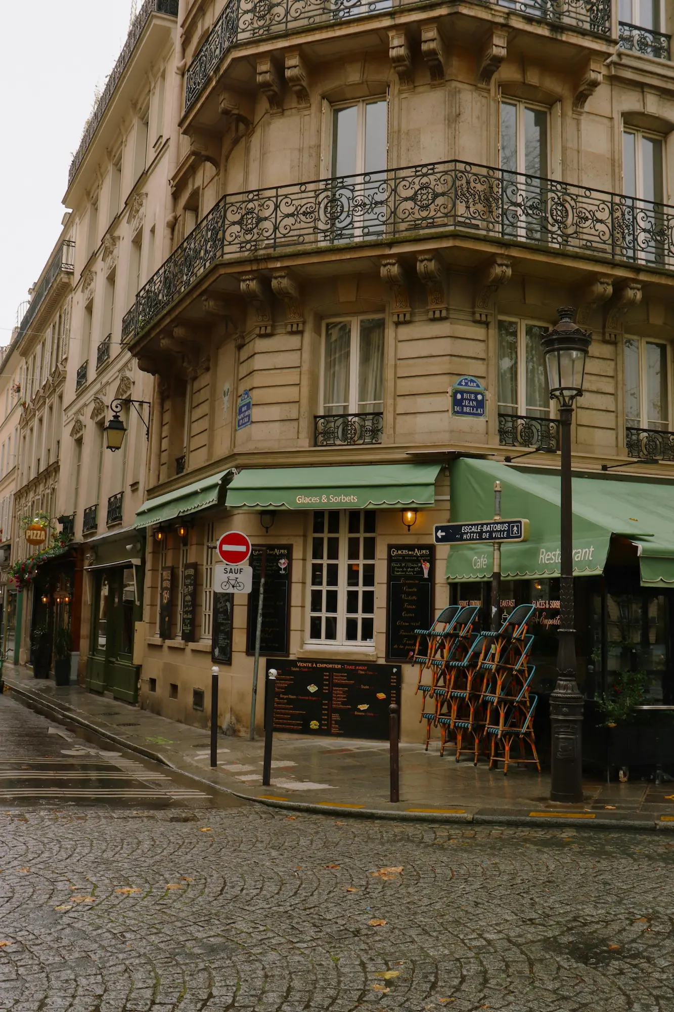 Façade d'un café-restaurant parisien avec un auvent vert, des chaises empilées et des rues pavées mouillées.