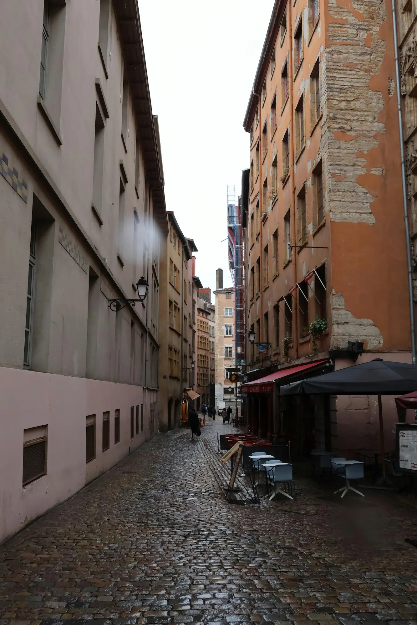 Ruelle pavée étroite bordée de vieux bâtiments avec des tables de café vides sous des parasols, sous un ciel gris.