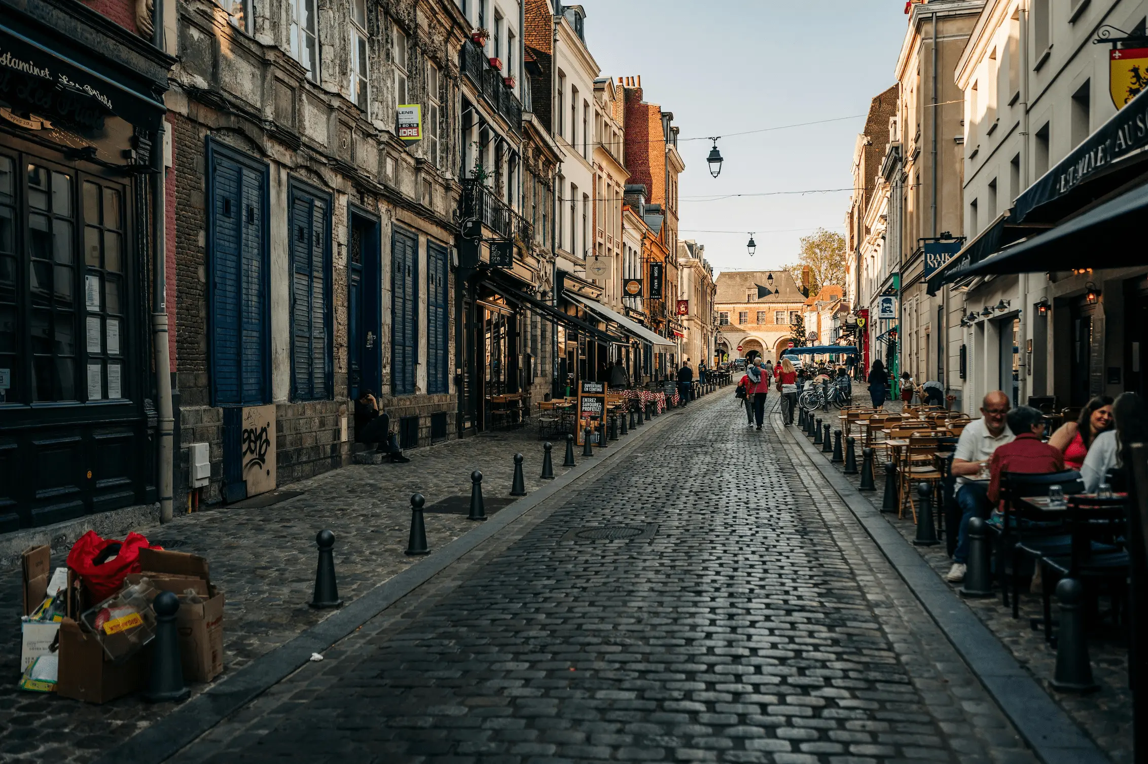 Rue pavée en ville avec des bâtiments anciens, des cafés en terrasse et des passants sous un ciel clair.