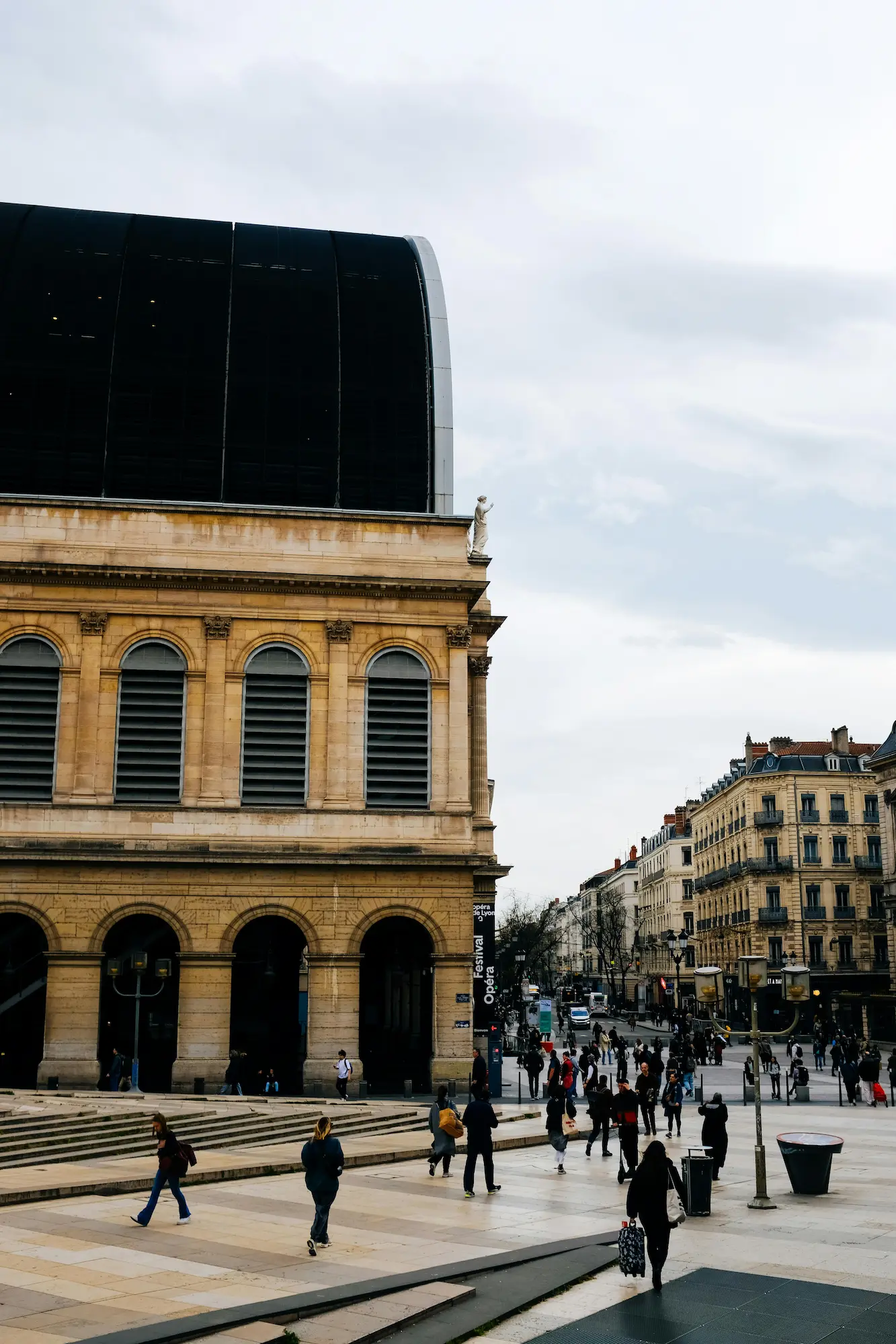 Place animée devant un bâtiment historique en pierre avec de grandes arches et des fenêtres voûtées, des personnes marchant dans une rue urbaine.