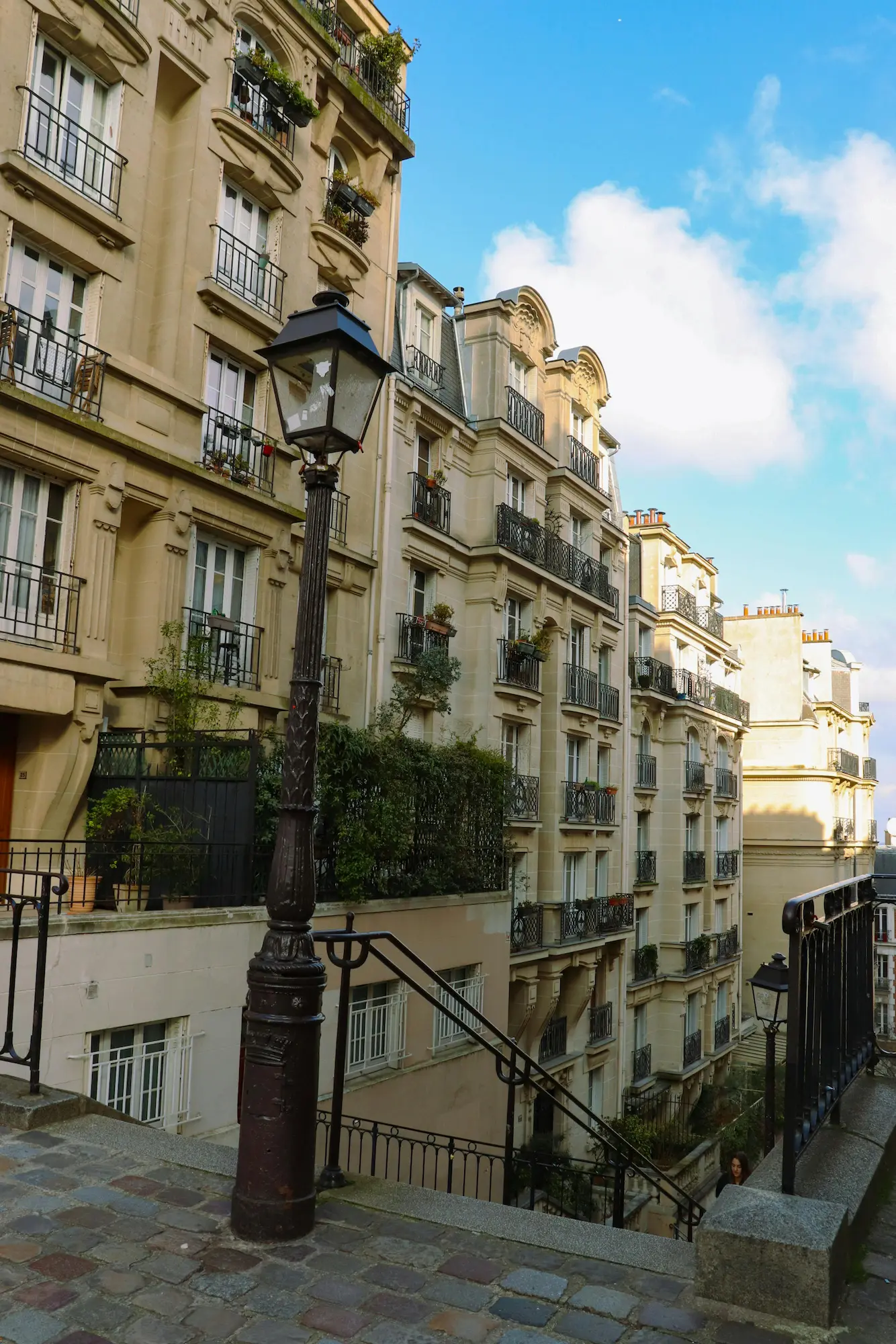 Rue pavée avec lampadaire ancien au premier plan et immeubles typiques avec balcons en fer forgé sous un ciel bleu partiellement nuageux.
