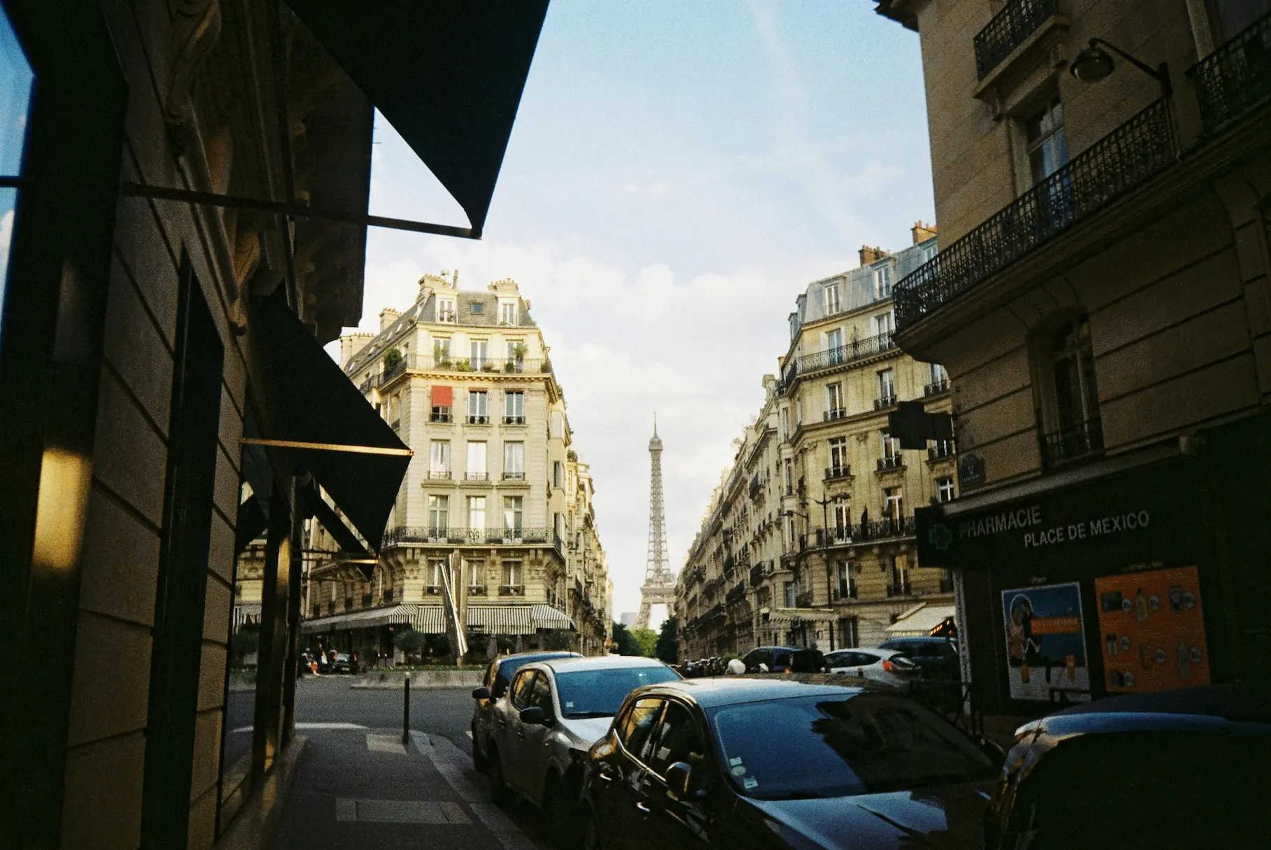 Rue parisienne avec des voitures en stationnement et la tour Eiffel visible au loin entre des immeubles haussmanniens.