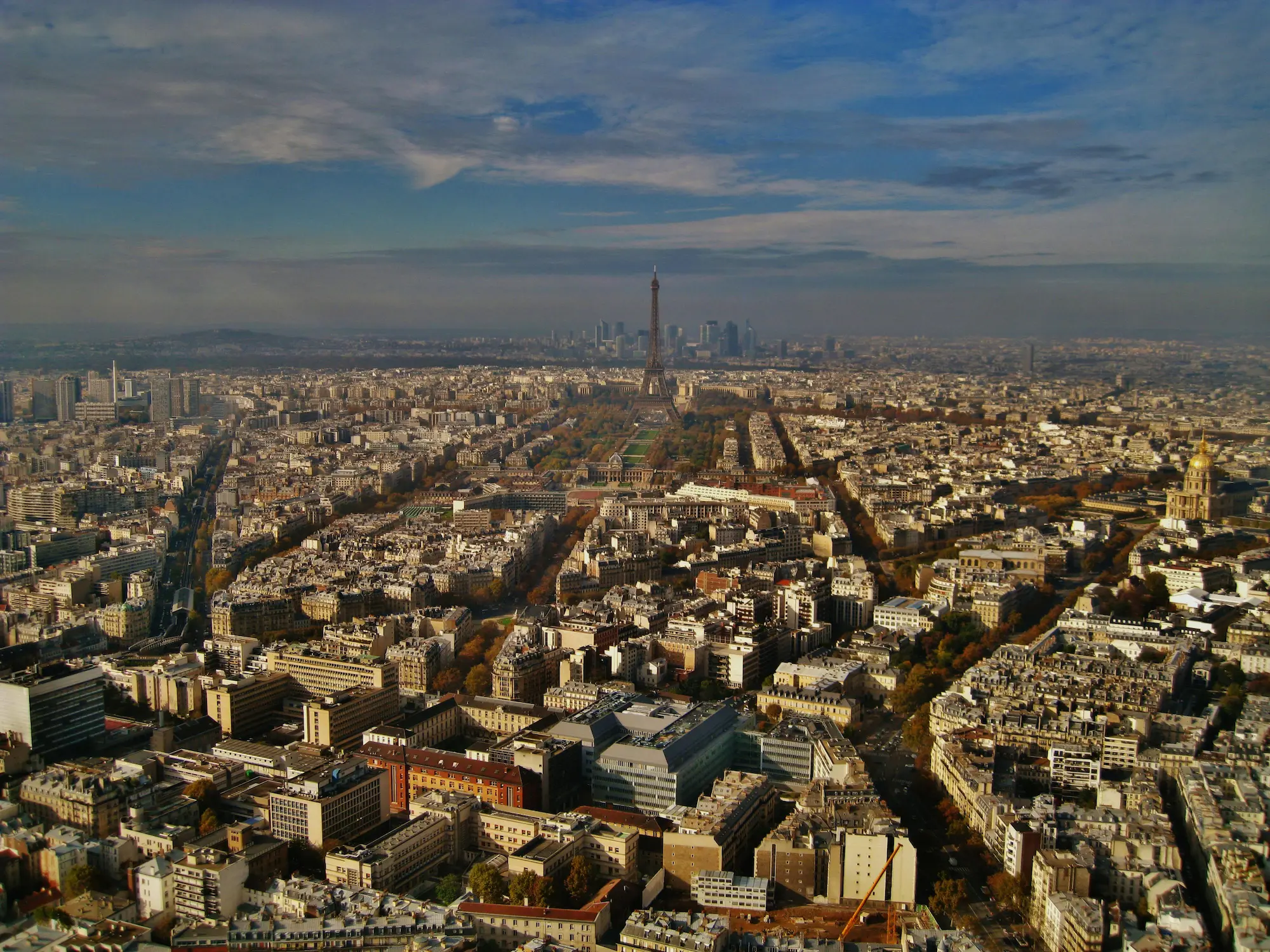 Vue aérienne de Paris avec la tour Eiffel au centre et des bâtiments urbains sous un ciel partiellement nuageux.