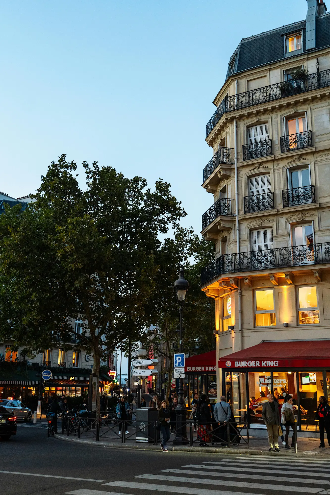 Personnes debout et marchant sur un trottoir à côté d'un Burger King dans un bâtiment haussmannien à Paris au crépuscule.