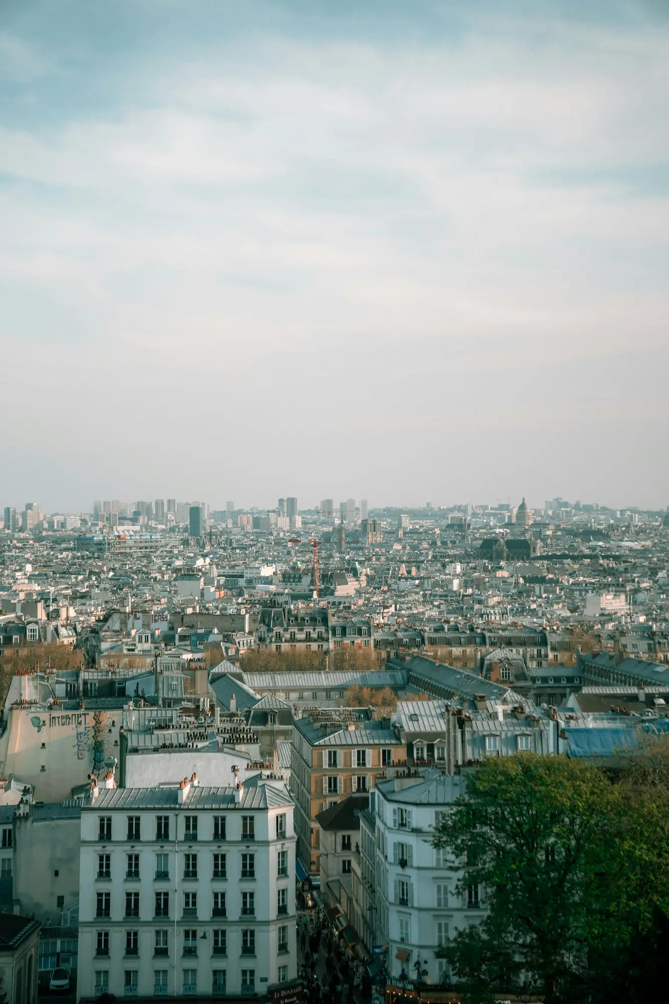 Vue aérienne d'une ville européenne dense avec des bâtiments historiques et des toits en zinc sous un ciel partiellement nuageux.