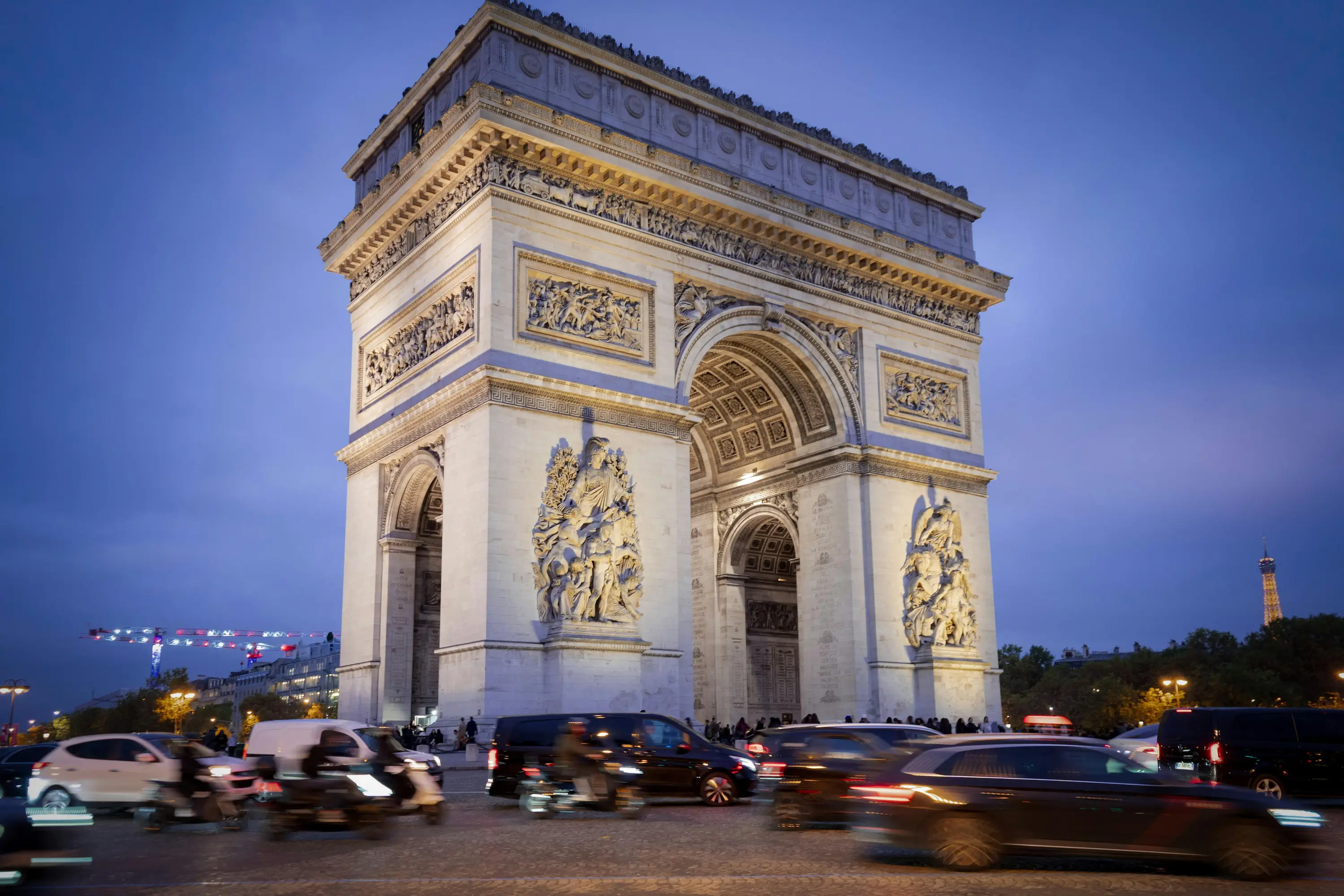 Vue en soirée de l'Arc de Triomphe à Paris avec des voitures et motos en mouvement autour.