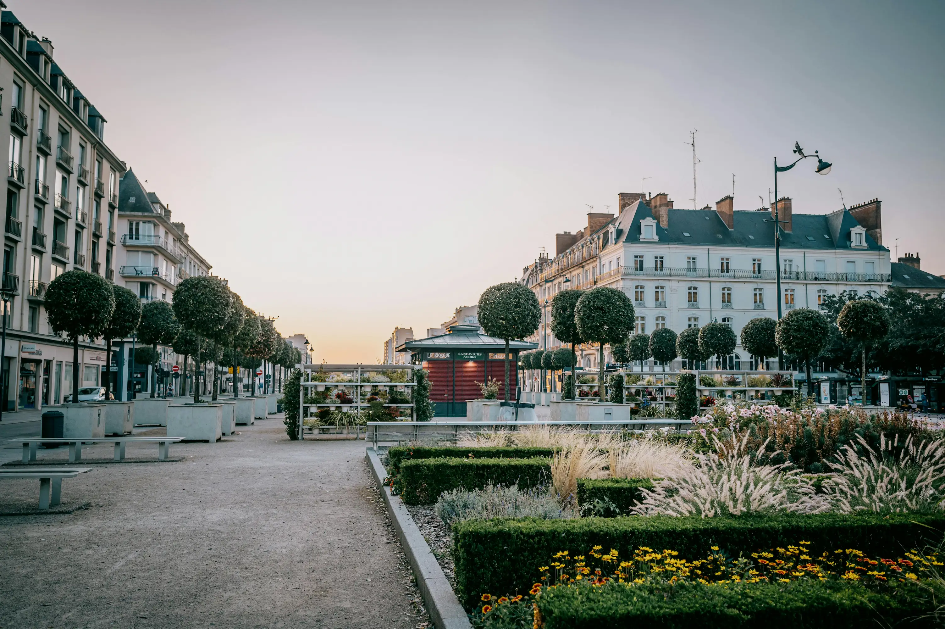 Place urbaine avec allée en gravier, bordée de bancs, de topiaires sphériques et de parterres fleuris devant des immeubles haussmanniens au coucher du soleil.