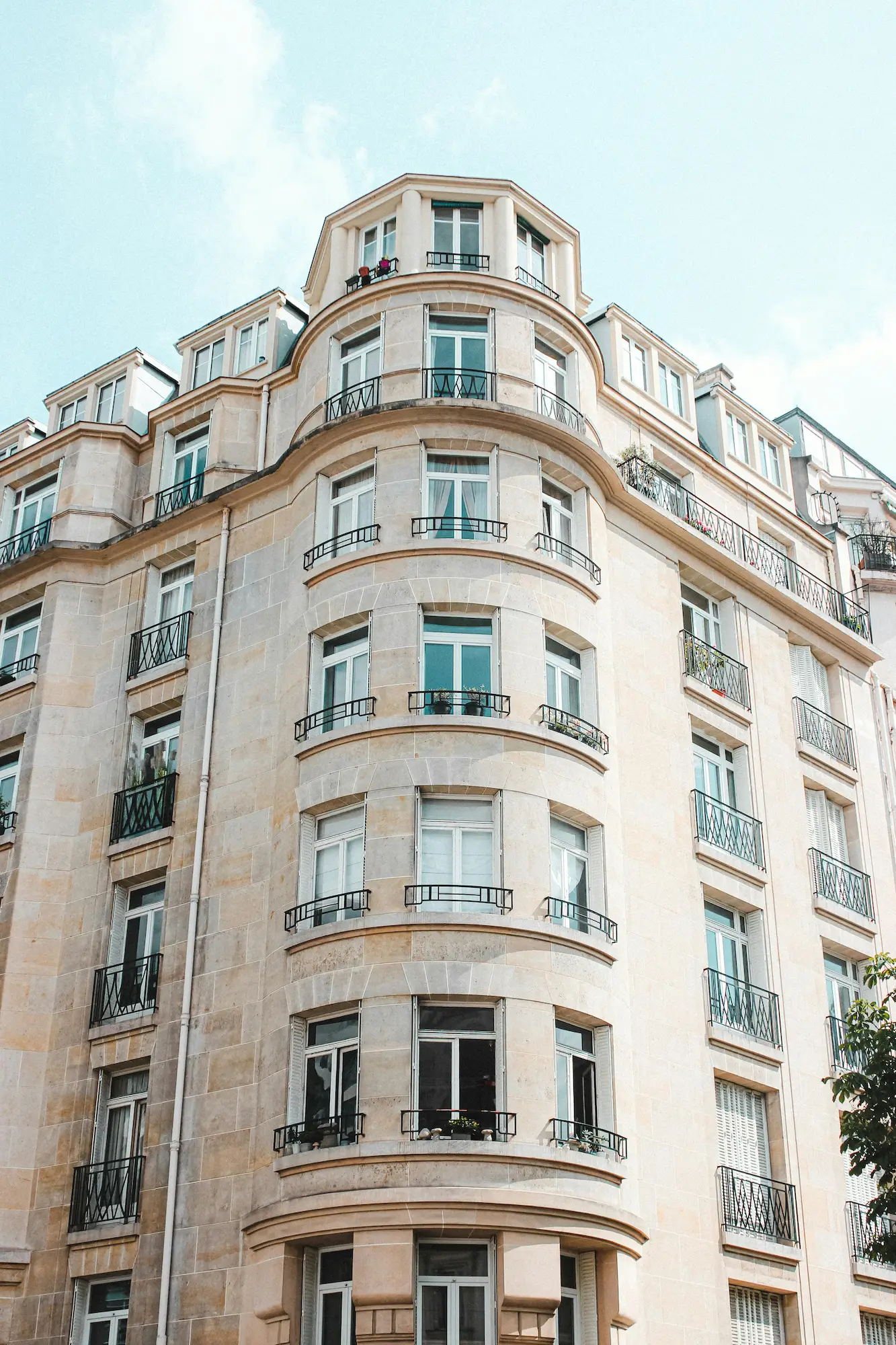 Façade d'un immeuble en pierre beige avec plusieurs fenêtres et balcons en fer forgé, sous un ciel bleu clair.