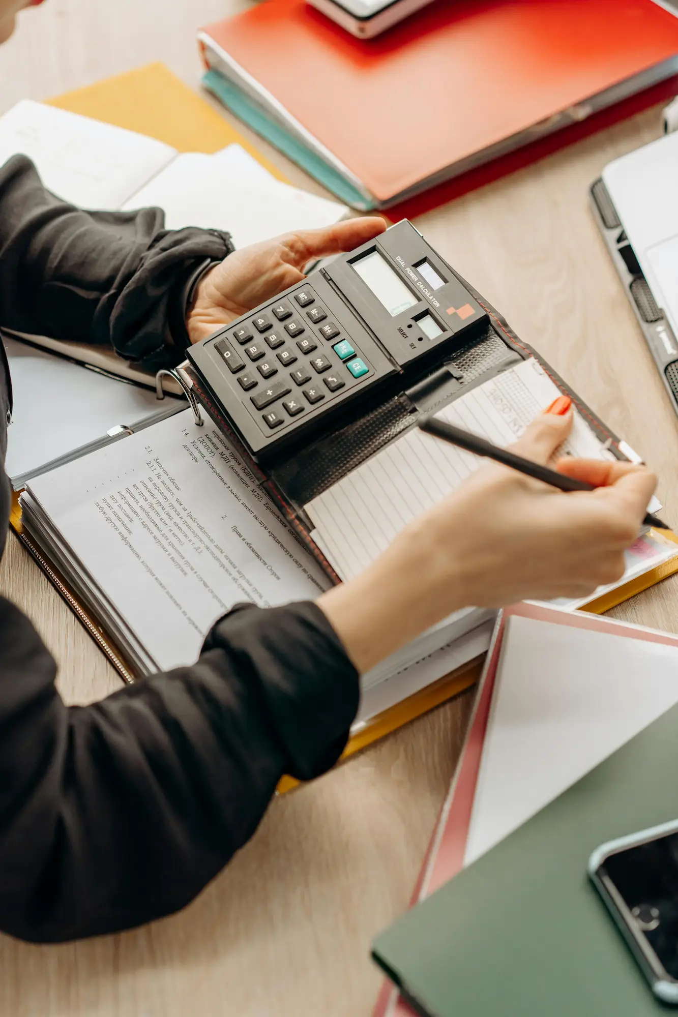Une personne utilise une calculatrice portable avec un carnet ouvert et prend des notes avec un crayon sur un bureau avec des dossiers et un téléphone portable.