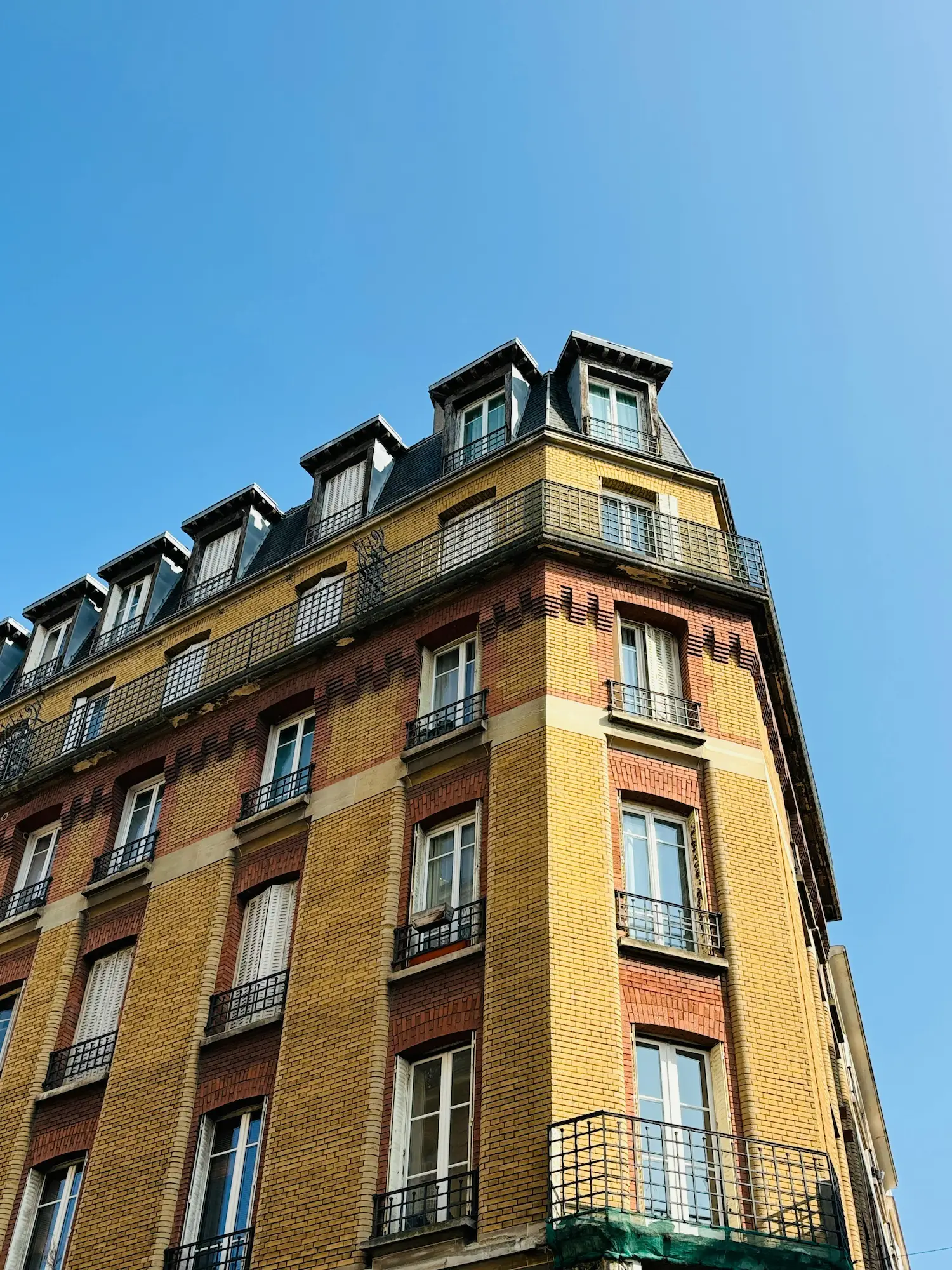 Bâtiment d'angle en briques jaunes et rouges avec balcons en fer forgé sous un ciel bleu clair.