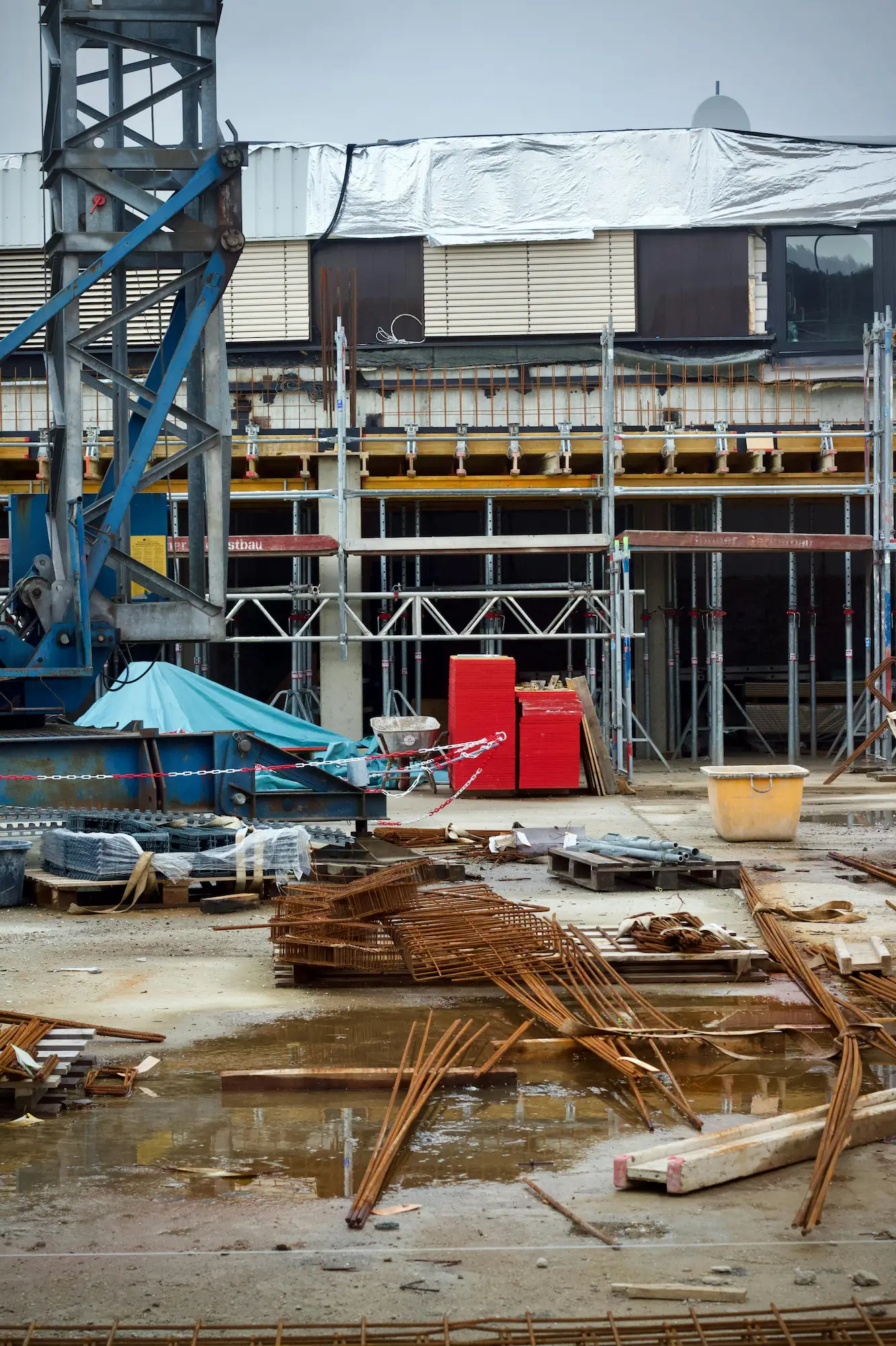 Chantier de construction avec échafaudages métalliques, matériaux de construction rouillés et flaque d'eau au sol.