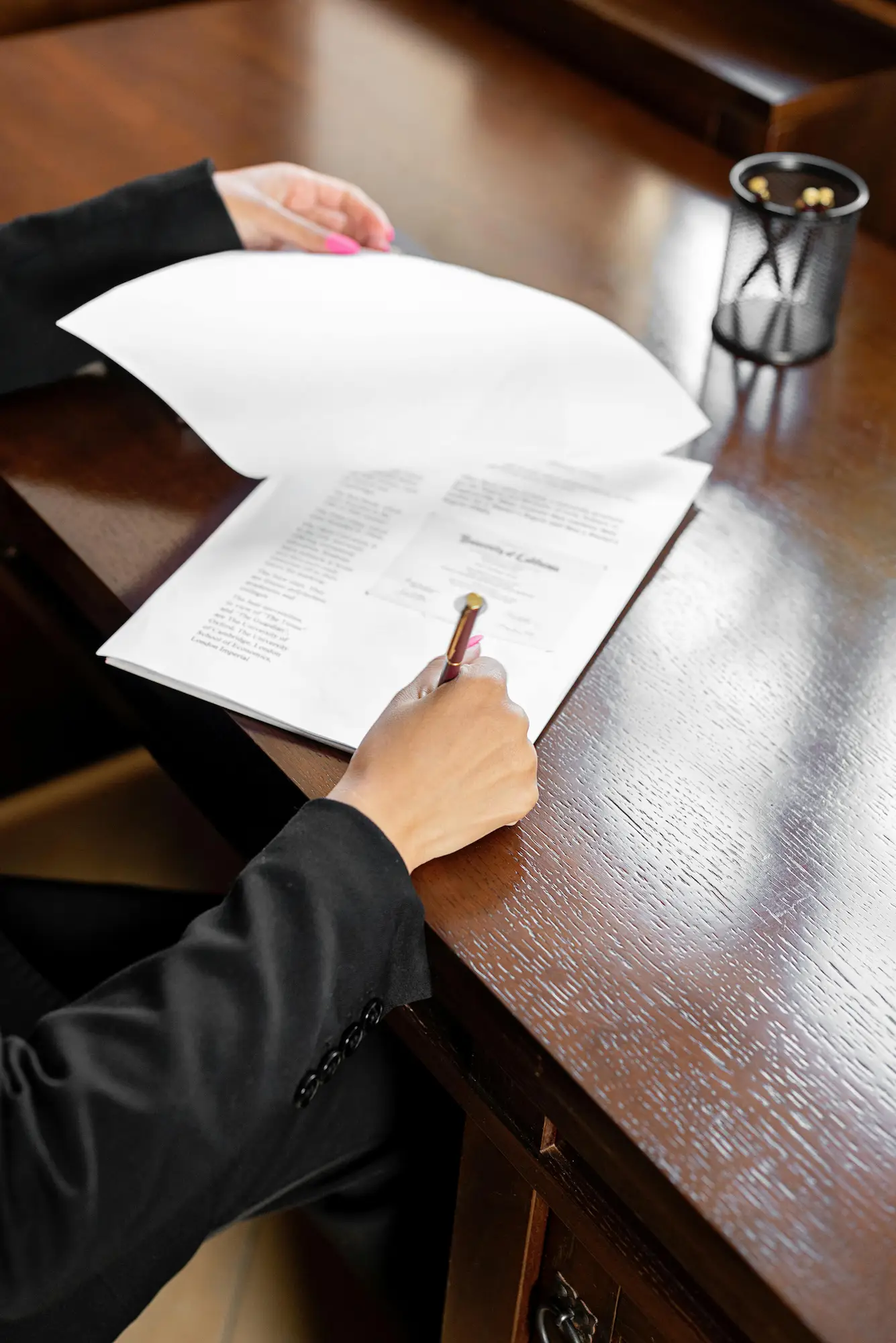 Person reviewing and signing documents on a wooden desk with a pen and a mesh pencil holder.