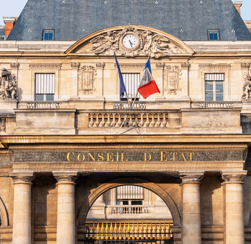 Façade du bâtiment du Conseil d'État avec deux colonnes, un horloge et drapeaux français.