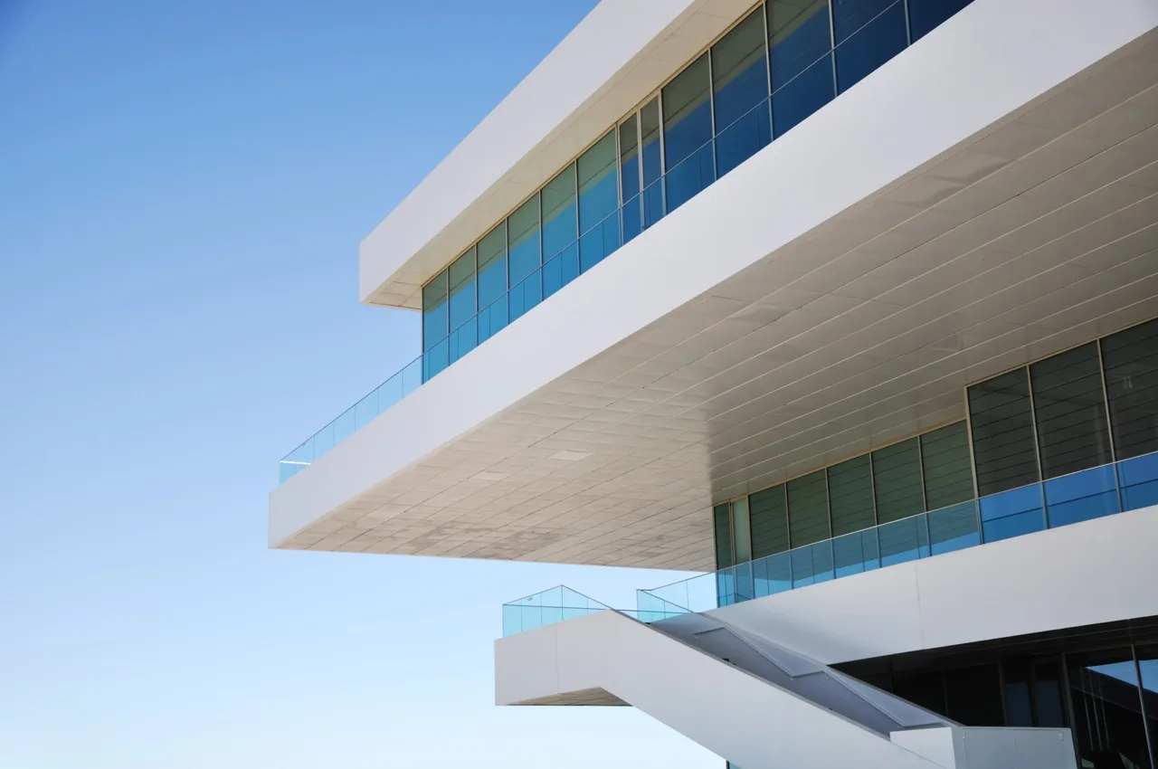 Bâtiment moderne blanc avec grandes fenêtres aux cadres bleus et escalier extérieur sous ciel bleu clair.
