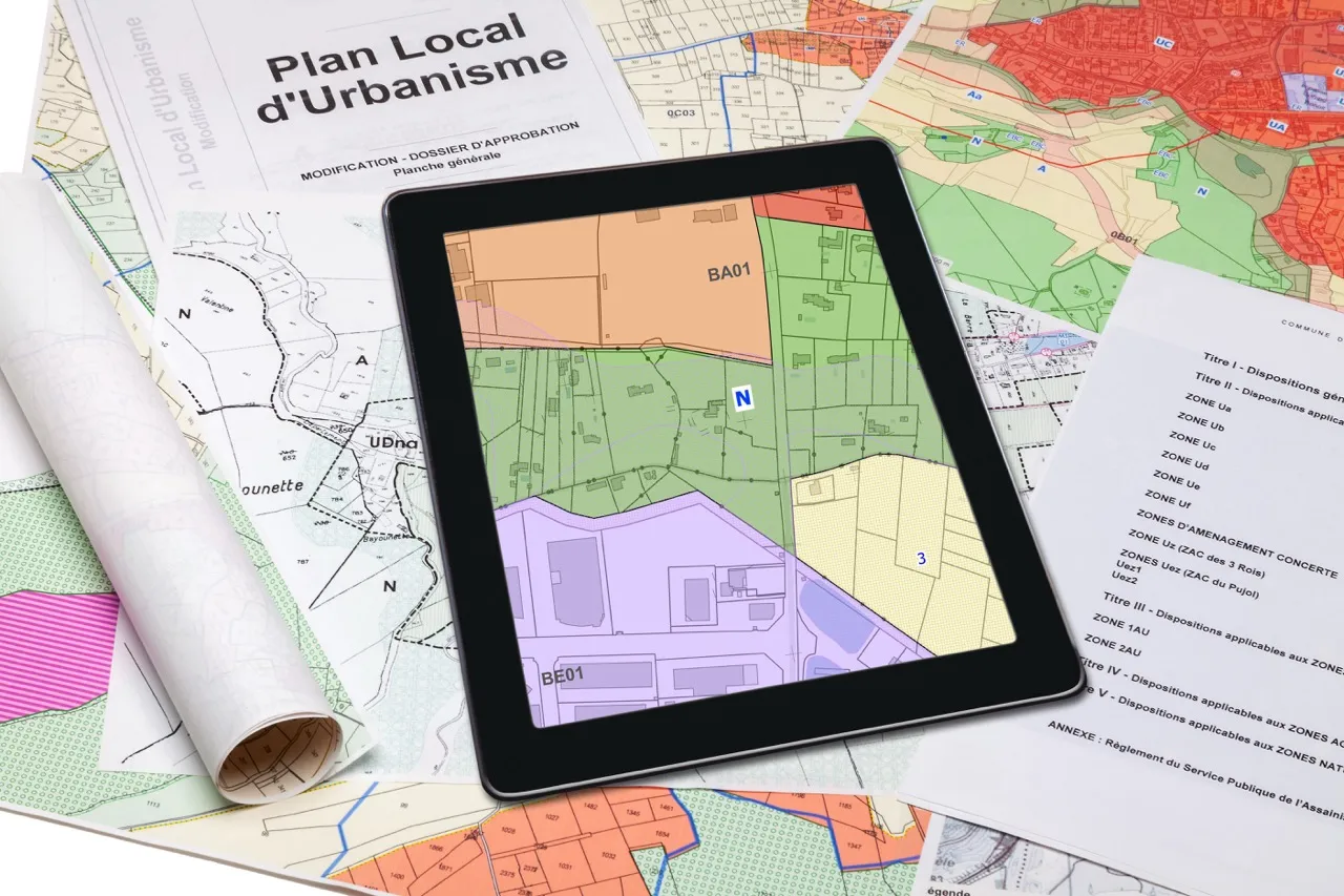 Plan local d'urbanisme avec des cartes papier et une tablette affichant des zones colorées pour l'aménagement.