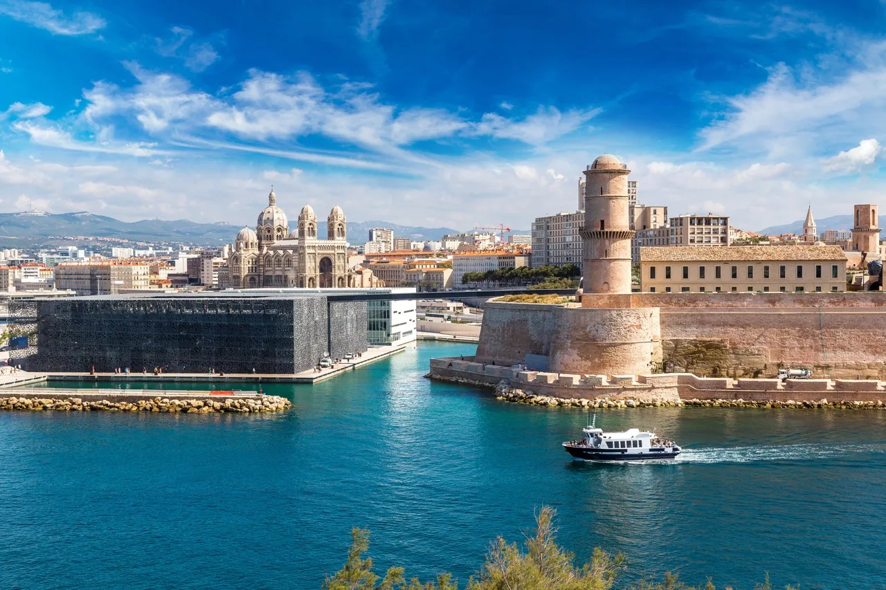 Vue du Vieux-Port de Marseille avec la cathédrale et le fort Saint-Jean sous un ciel bleu clair.