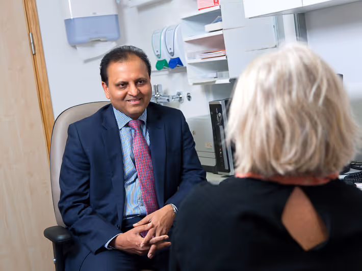 Male doctor in suit smiling and talking to a seated woman with white hair in a medical office.
