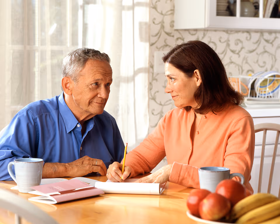 Elderly man and woman sitting at a table, holding a pencil and notebook, sharing a moment.