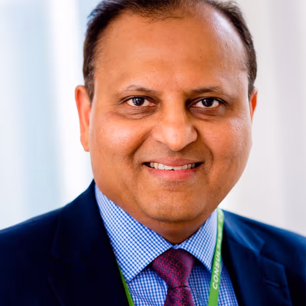 Smiling man wearing a dark suit, checkered shirt, red patterned tie, and green conference lanyard.