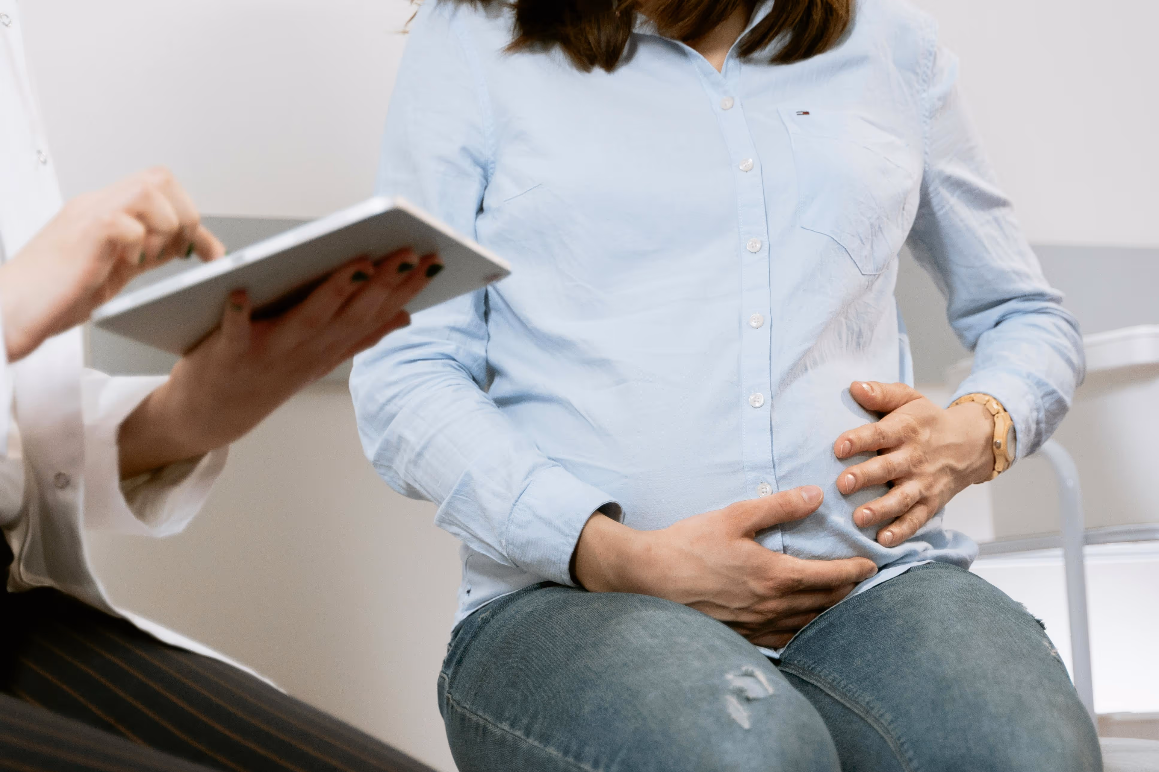 Pregnant woman in a blue shirt holding her belly during a medical consultation with a doctor.