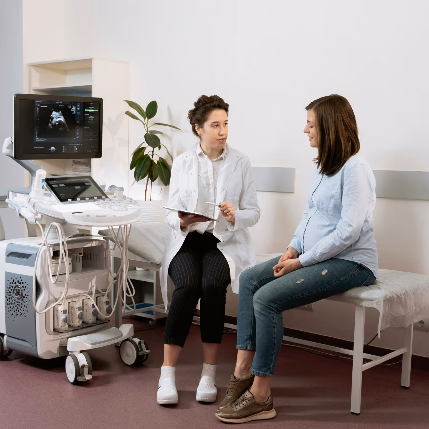 Pregnant woman consulting a female doctor with ultrasound equipment in a medical exam room.