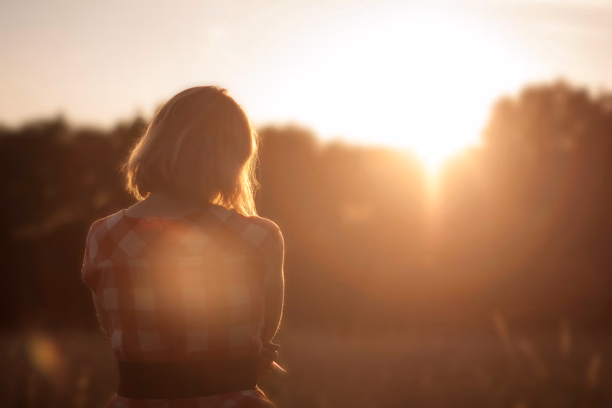 Woman in a checkered dress looking at a bright sunset in a natural outdoor setting.