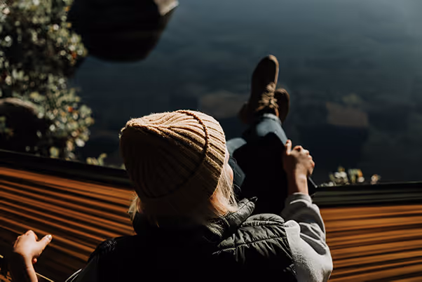 Person wearing a knit hat and jacket relaxing in a hammock above water and foliage.