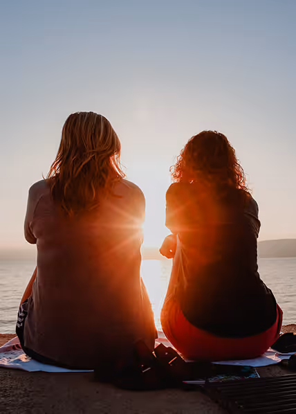 Two women sitting on a beach towel watching the sunset over the water.