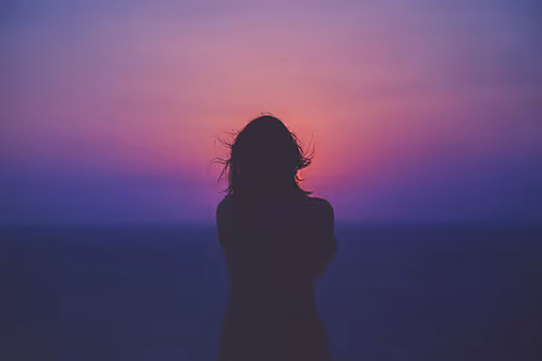 Silhouette of a person with windblown hair facing a vibrant sunset over the ocean.