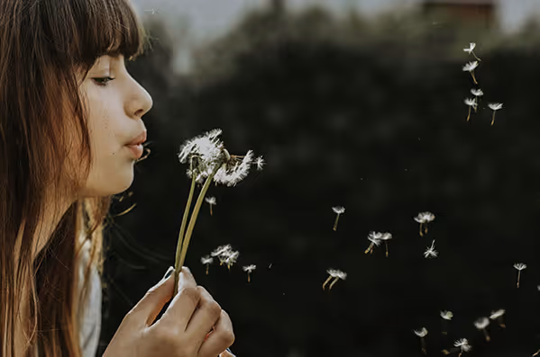 Young woman blowing dandelion seeds into the air outdoors with blurred dark green background.