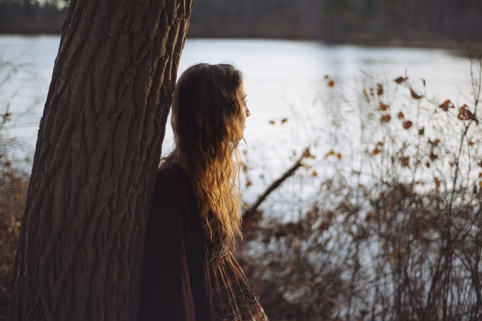 Woman with long hair leaning against a tree, looking out over a calm lake with dry foliage.