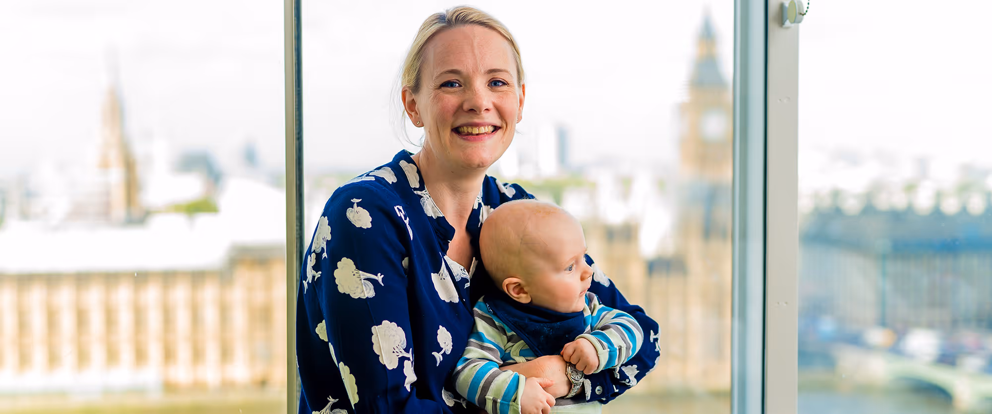 Smiling woman holding baby in front of a large window overlooking London cityscape with Big Ben.
