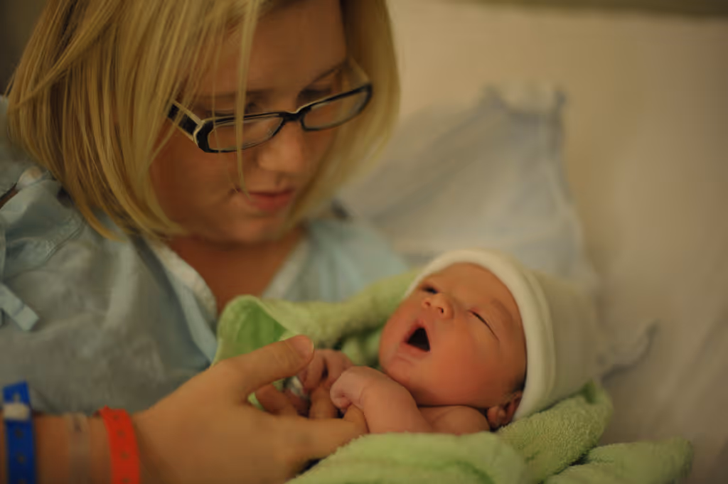 Newborn baby wrapped in green blanket wearing white hat held by woman in hospital gown and glasses.
