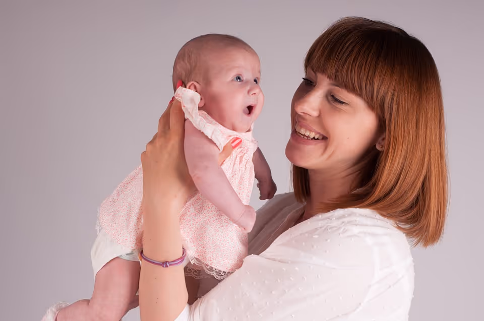 Smiling woman with red hair holding a surprised baby in a pink dress against a plain background.