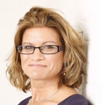 Smiling woman with glasses and blonde hair wearing a necklace and dark top against a white background.