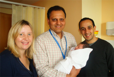 Three adults smiling, one holding a newborn baby wrapped in a white blanket in a hospital room.